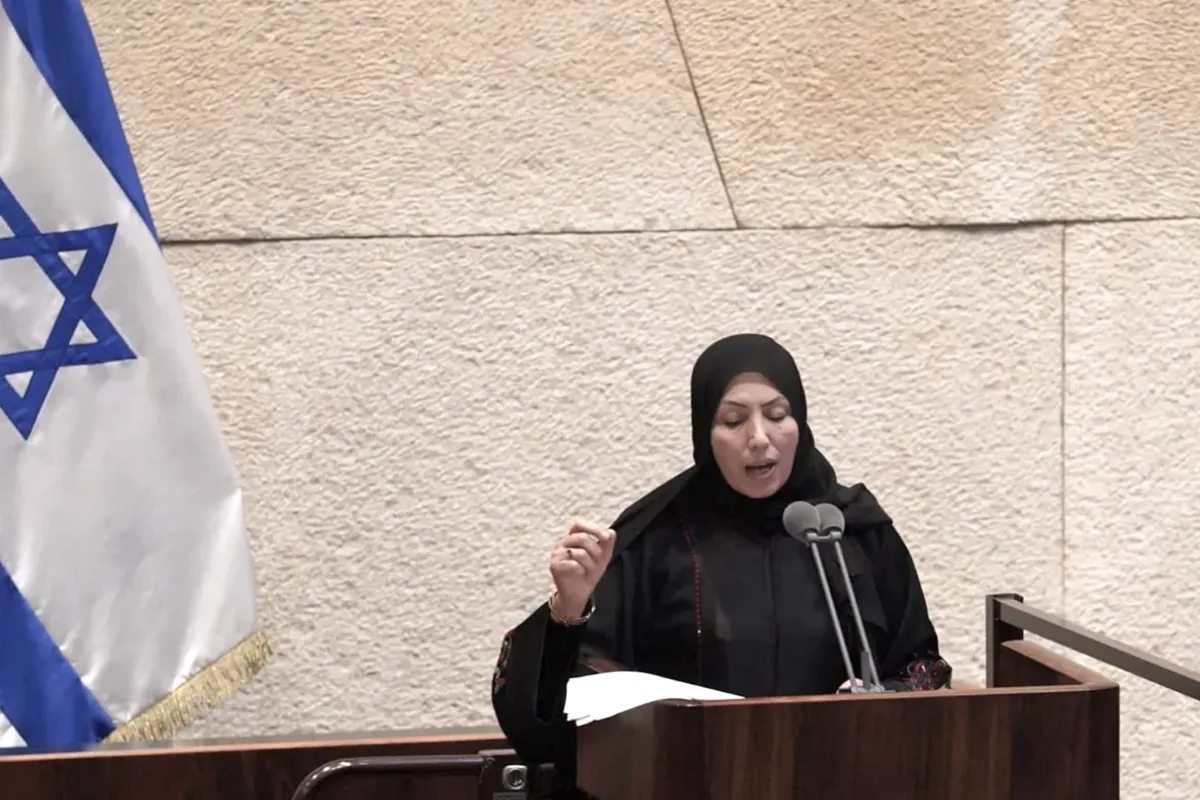 A woman in a black hijab delivers a speech at the podium of the Israeli Knesset, standing next to a large Israeli flag.