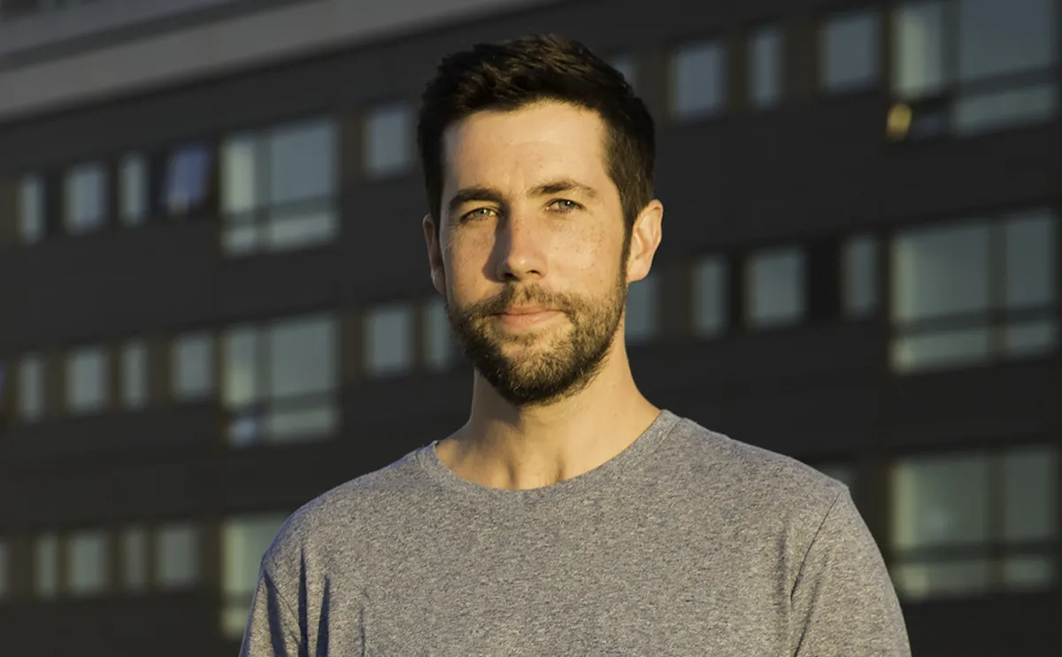 white man in gray shirt with beard over blurred building background