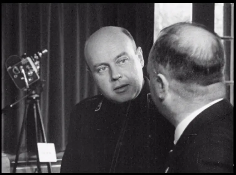 A bald white man, in a dark uniform, talks to another man with his back facing the camera.