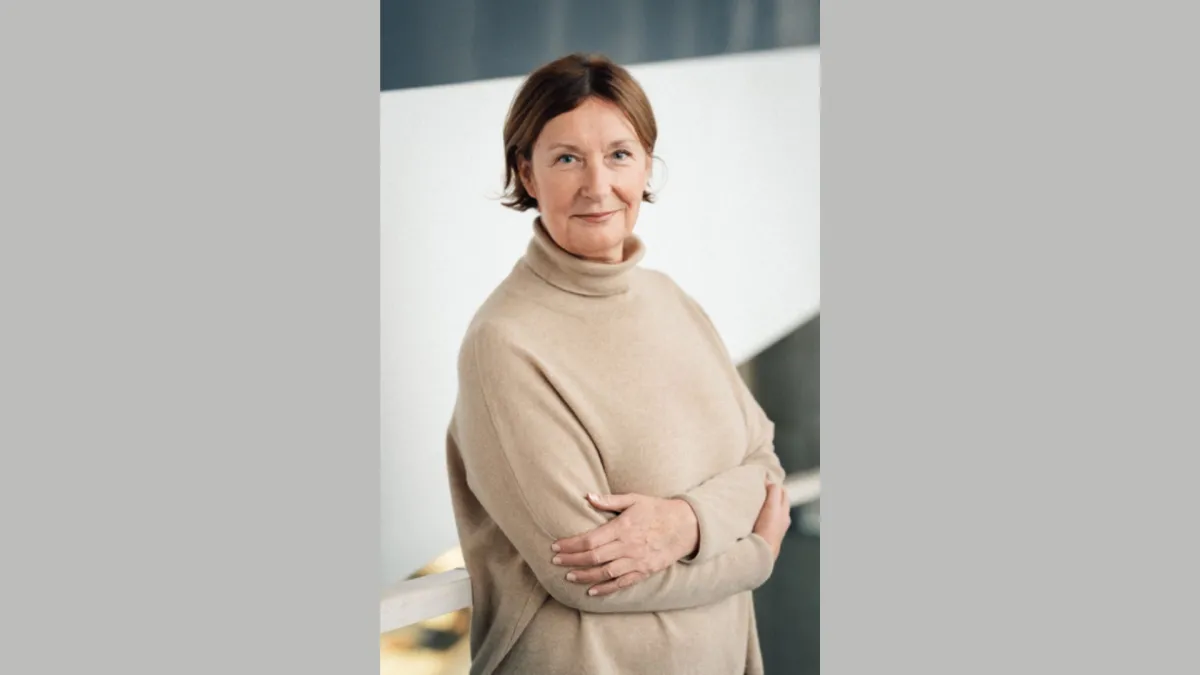 A light-skinned woman with cropped brown hair and a beige sweater gently smiles at the camera.