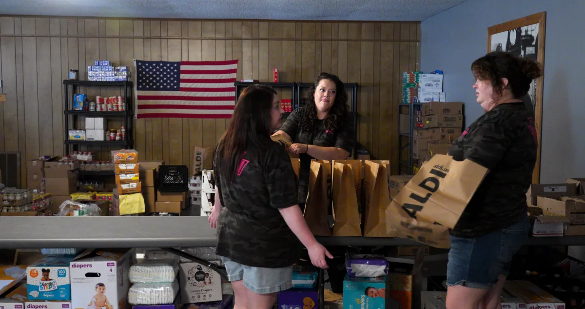 Three individuals carrying items in a pantry, an American flag in the background