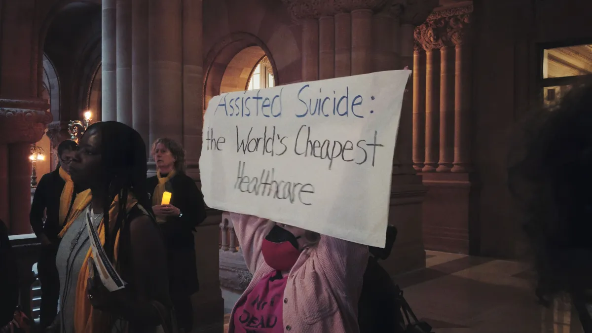 A woman in a pink jacket holds up a sign that reads: "Assisted Suicide: The World's Cheapest Healthcare."