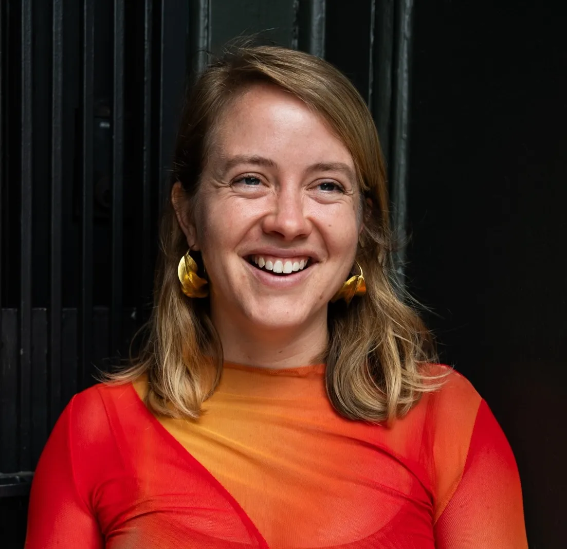 Headshot of an adult woman with light skin tone, blonde shoulder-length hair wearing a red and orage long-sleeved shirt and gold hoop earrings.