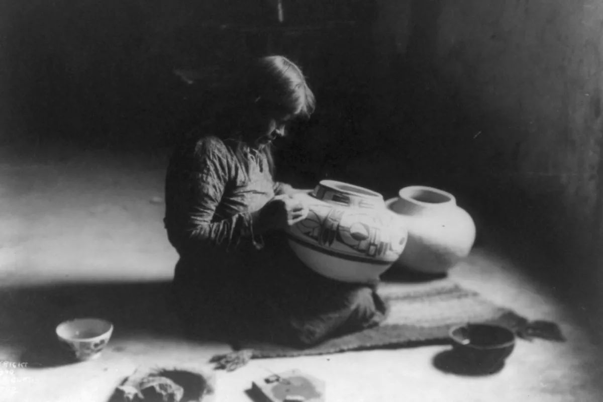 Old black & white photograph of a person sitting on the ground decorating pottery