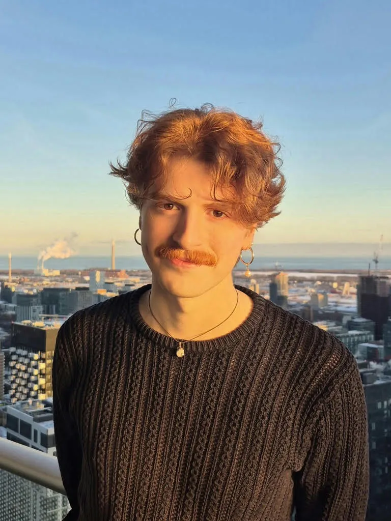 A red-head man with a mustache stands in front of a cityscape at sunset, looking at the camera.