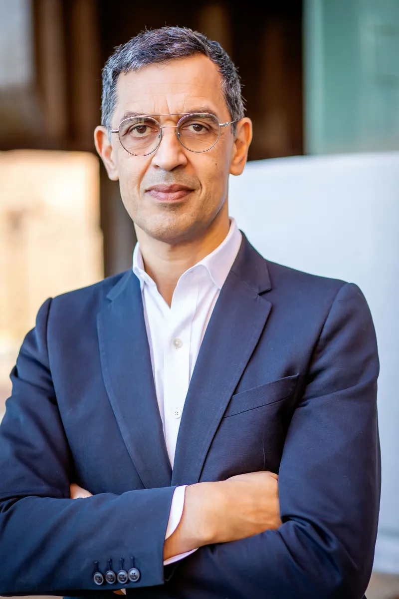 A man in glasses and a blue suit looks at the camera with his arms crossed.