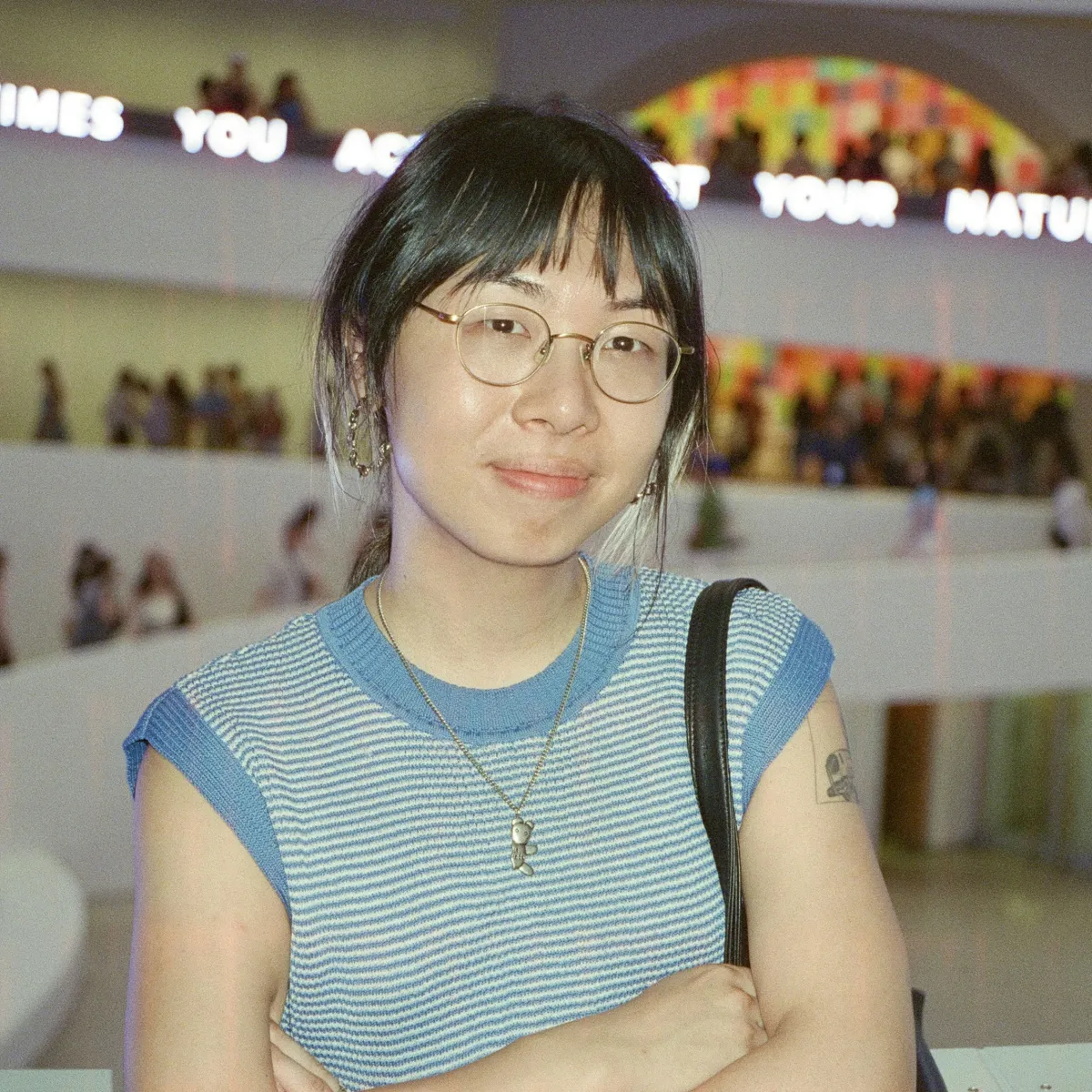 An asian woman with glasses stands looking at the camera with her arms crossed. She is wearing a blue T-Shirt and has her hair in a ponytail.