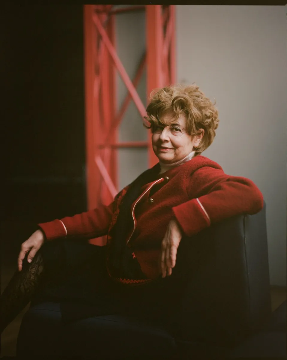 An older woman in a red skirt suit sits in a chair with her arm across the back, turning her head from the side to face the camera.