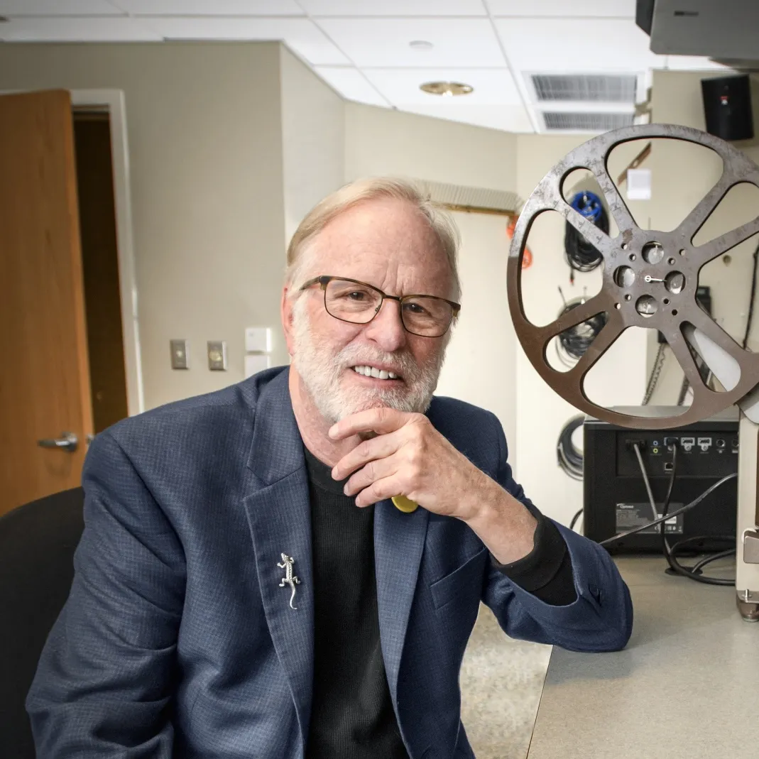 In front of a film projector, a white man wearing glasses and with a white beard sits at the booth.