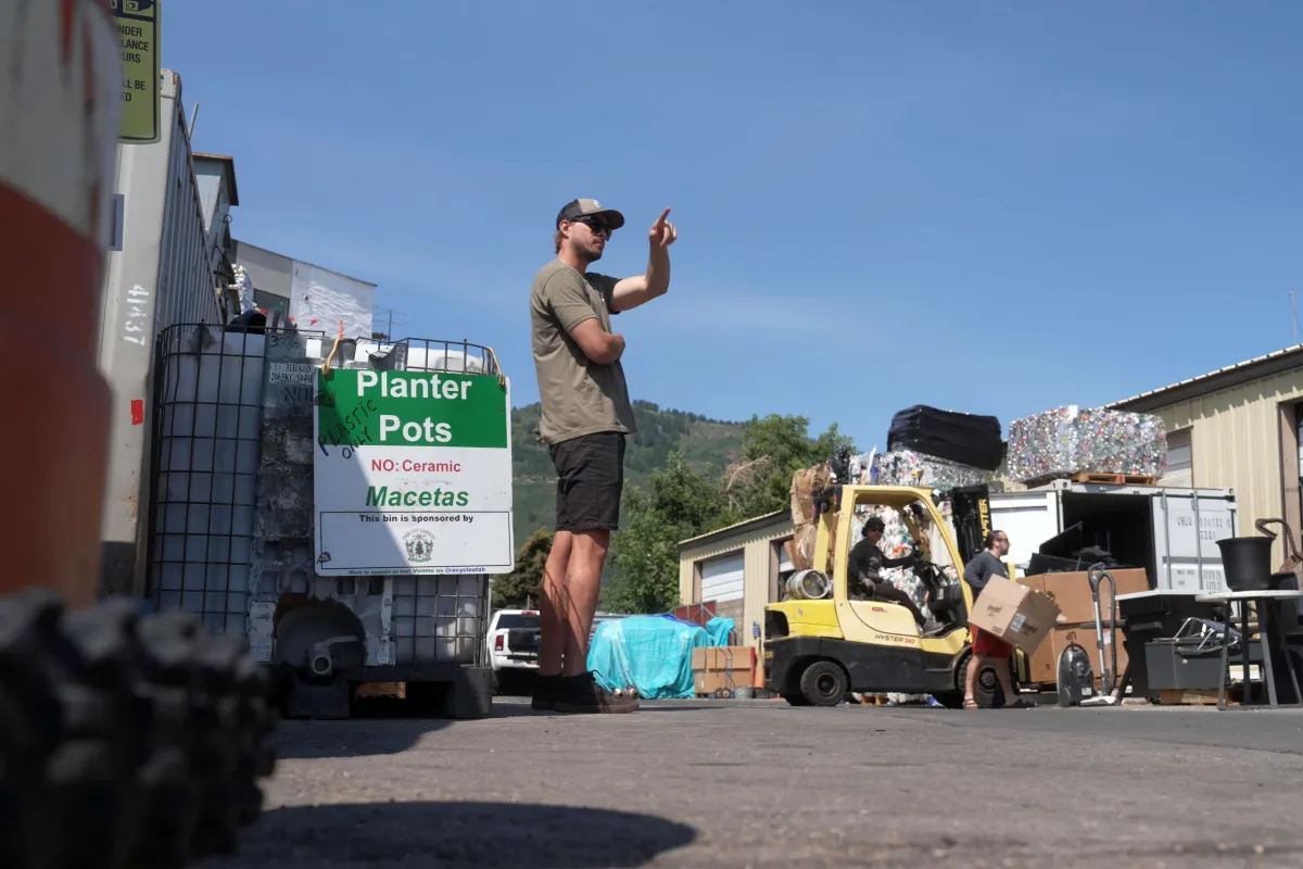 A man in a cap and sunglasses stands near a bin labeled “Planter Pots,” pointing into the distance. Behind him, a forklift carries crushed recyclables as two workers load boxes near a shipping container. The scene is set against a backdrop of trees, hills and a clear blue sky.