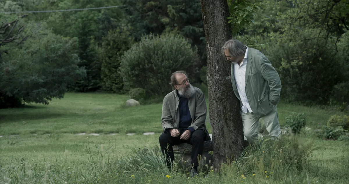 In a lush green field, two men, one sitting and one standing, talk to each other.
