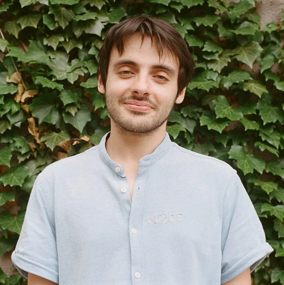 A brunette white man stands in front of a wall covered in ivy and looks at the camera.