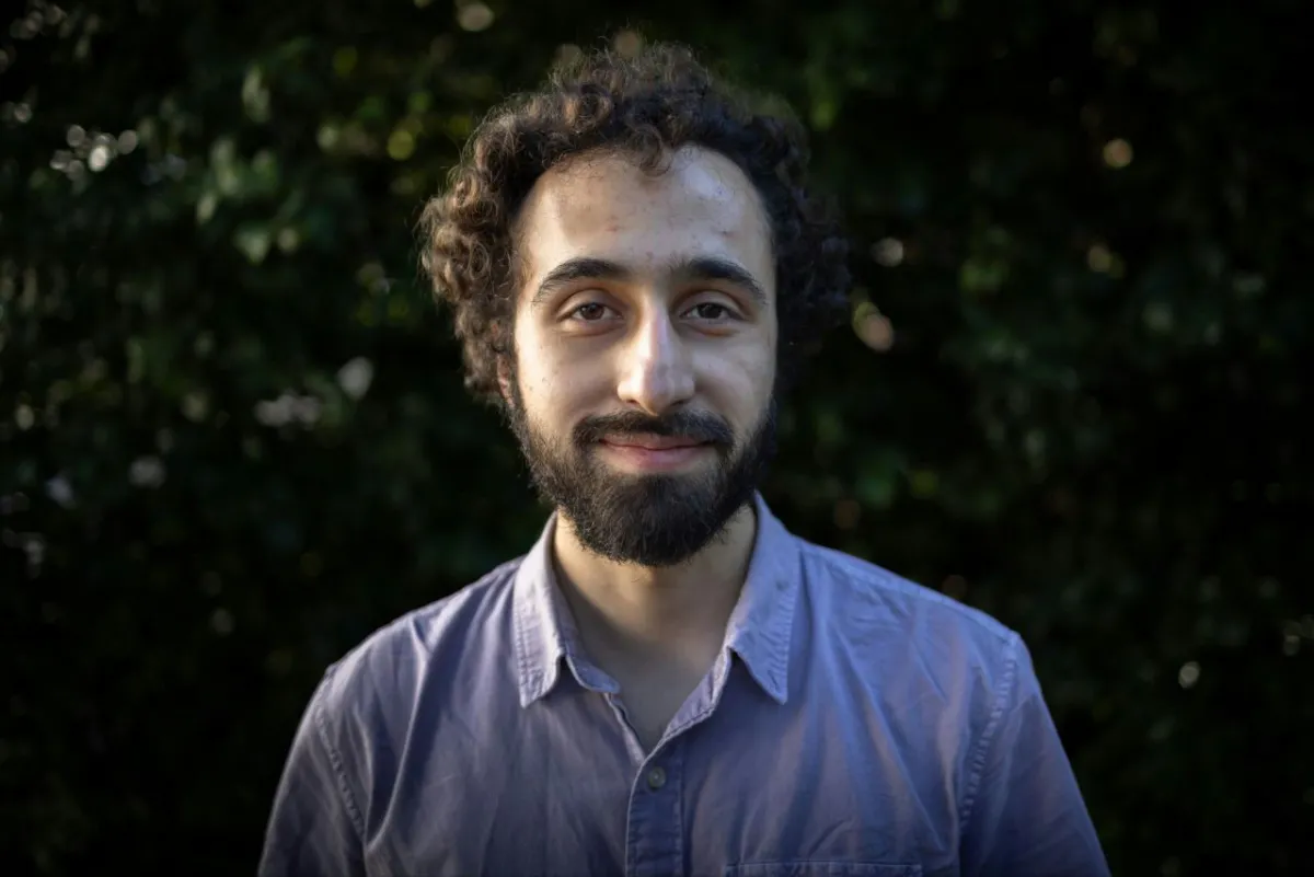 A man with a beard and blue shirt stands looking at the camera and smiles with the sun illuminating half his face. 