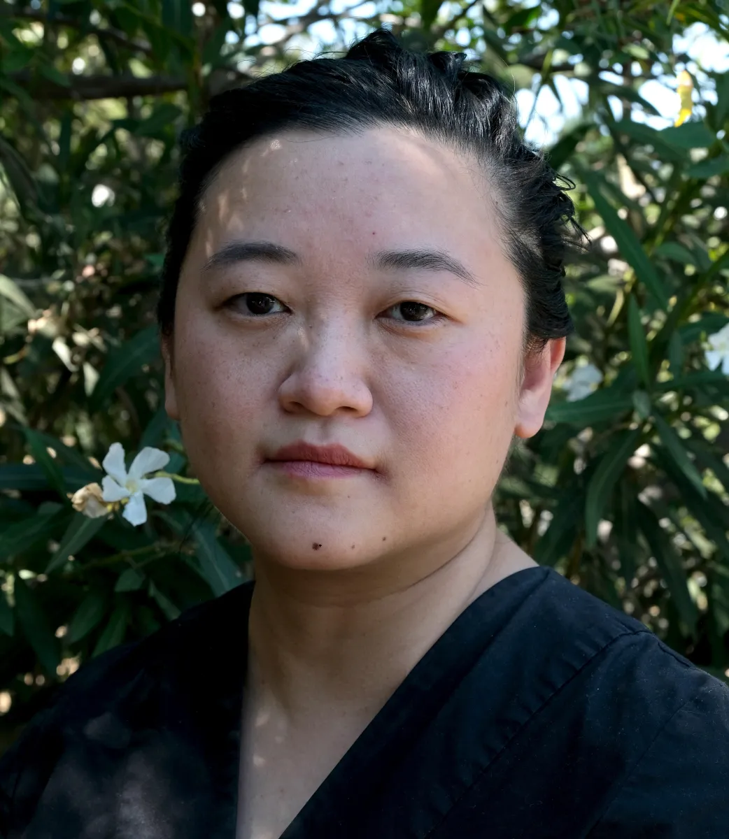 Portrait of Abby Sun, IDA's director of programs and editor of Documentary magazine. An Asian woman with short hair, wearing a black blouse and standing in front of a white flower bush.
