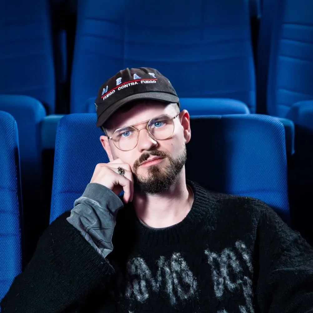 A white man wearing glasses and a thick black sweater sits in the middle of a theater with blue seats.