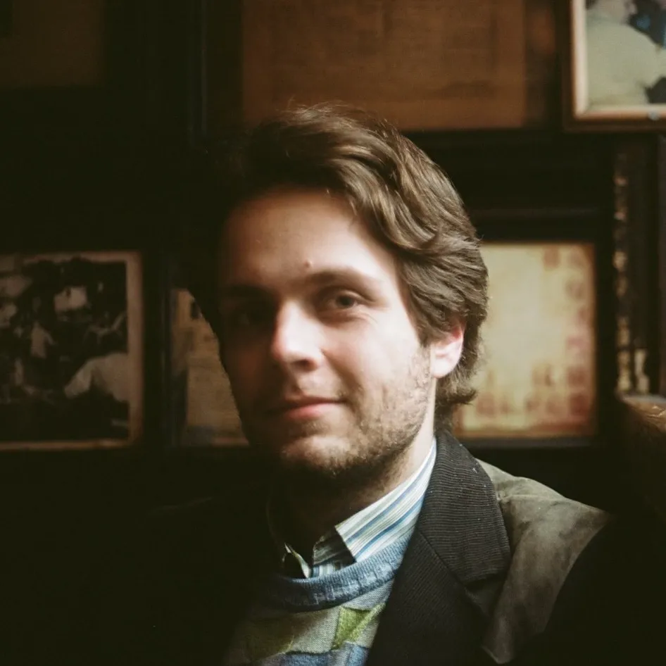 A man with wavy brown hair sits in a darkened room.