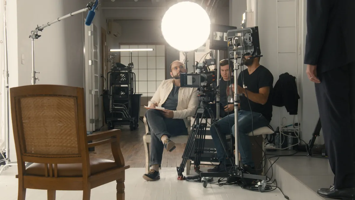 In a white studio, a white man wearing a white jacket sits in a chair, behind a camera and lights.