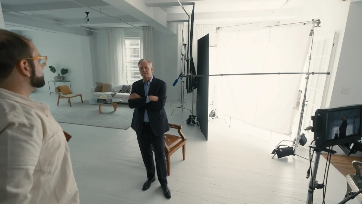 In a studio, Chris Hansen stands with his arms crossed, looking at a bearded white man in a white jacket.