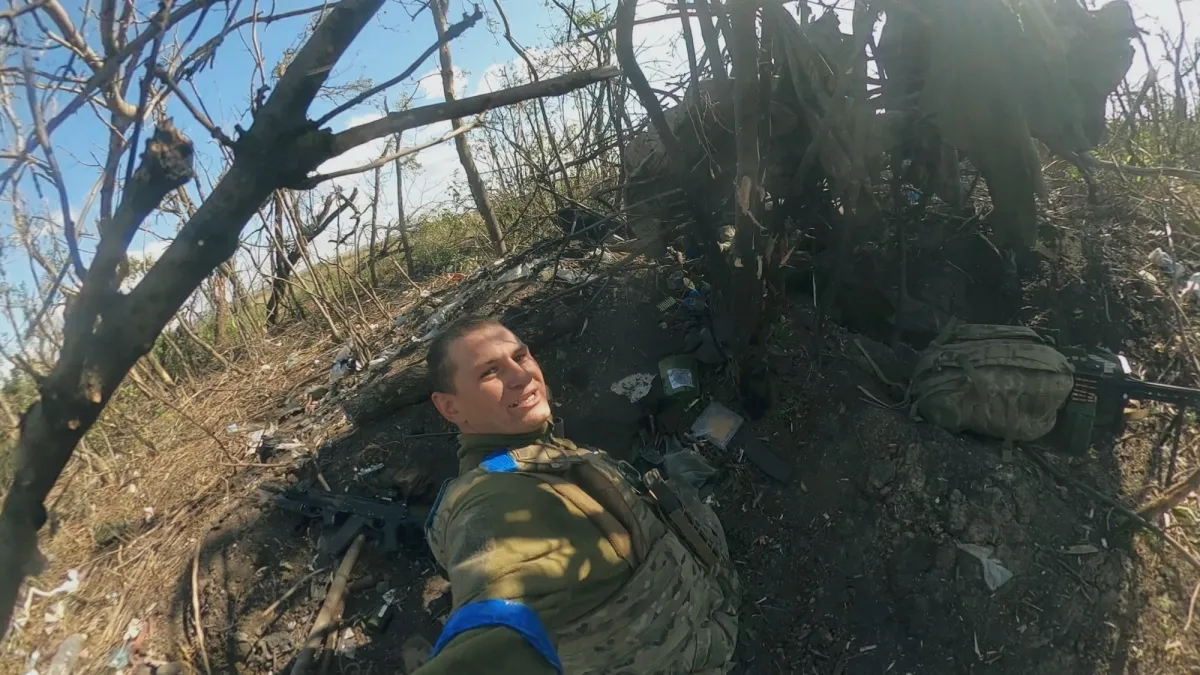 A soldier in a field takes a selfie that shows a makeshift campground around him