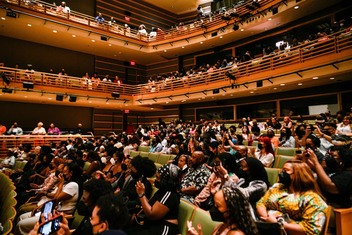 A crowded auditorium with green seats full of a clapping, sitting audience.