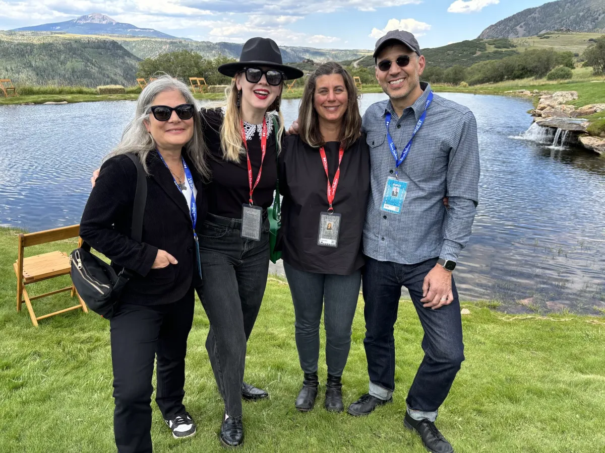 On the grassy banks of a lake, four white people wearing festival lanyards stand casually and happily.