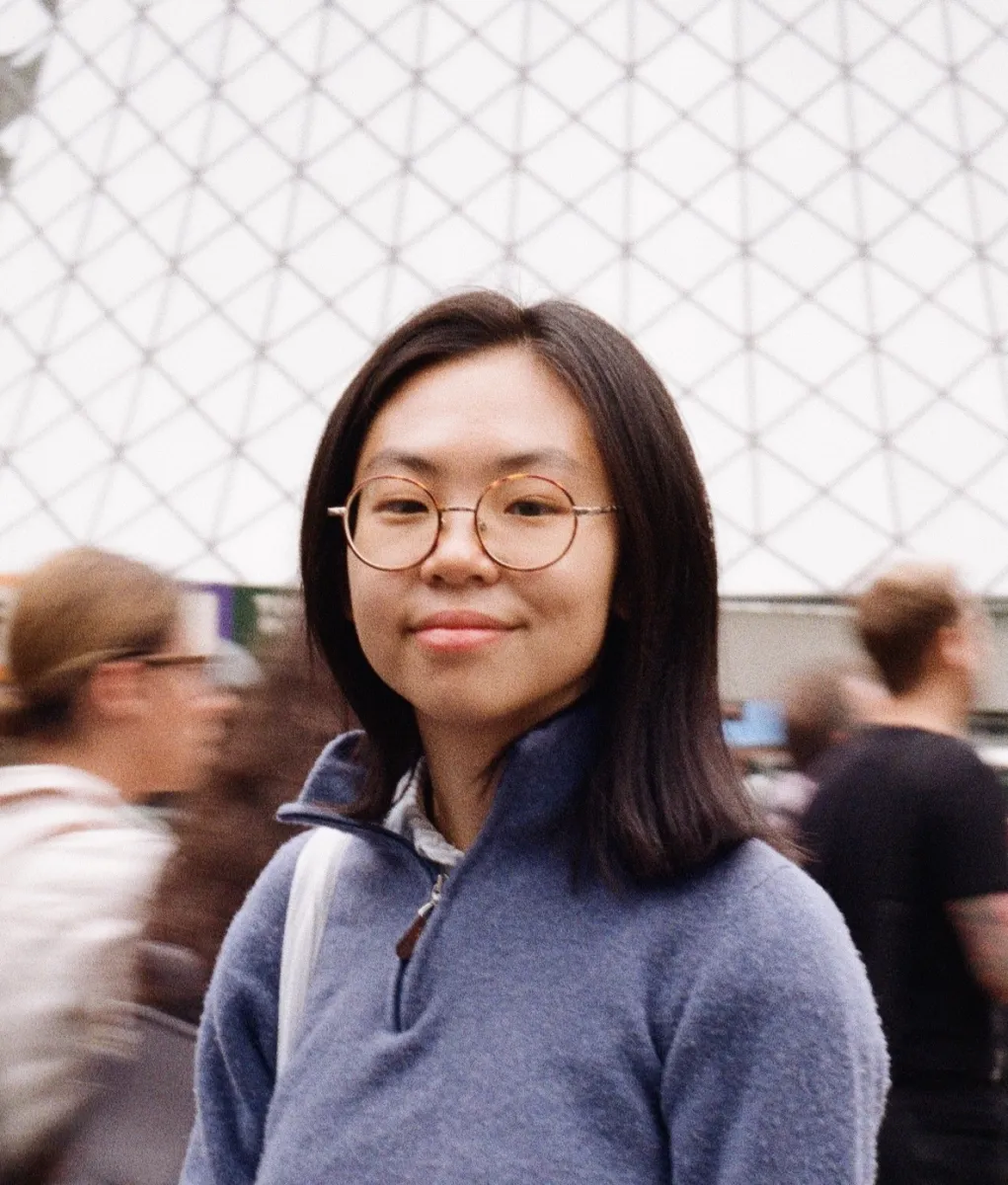 Headshot of a person with shoulder-length black hair standing outside in a plaza.