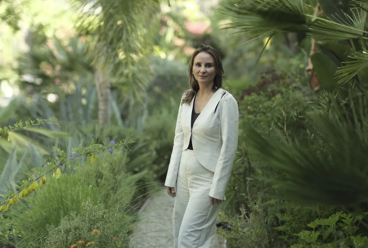 Woman with shoulder length brown hair in a white suit stands against a lush green garden backdrop.