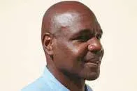A bald Black man seen in close-up wearing a blue shirt against a light background