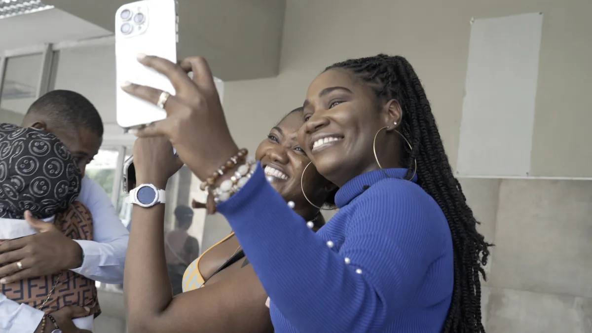 Vanessa and Tamara stand close together, smiling brightly as they take a selfie on a smartphone. Vanessa, in a royal blue top with long braids and hoop earrings, holds the phone out in front of them, while Tamara, dressed in a patterned outfit, leans in beside her. Both women radiate joy, their wide smiles filling the frame. Just behind them, Vanessa’s husband embraces someone tightly, his face pressed against a shoulder. The moment is tender yet bittersweet—Vanessa and Tamara are capturing one last photo t