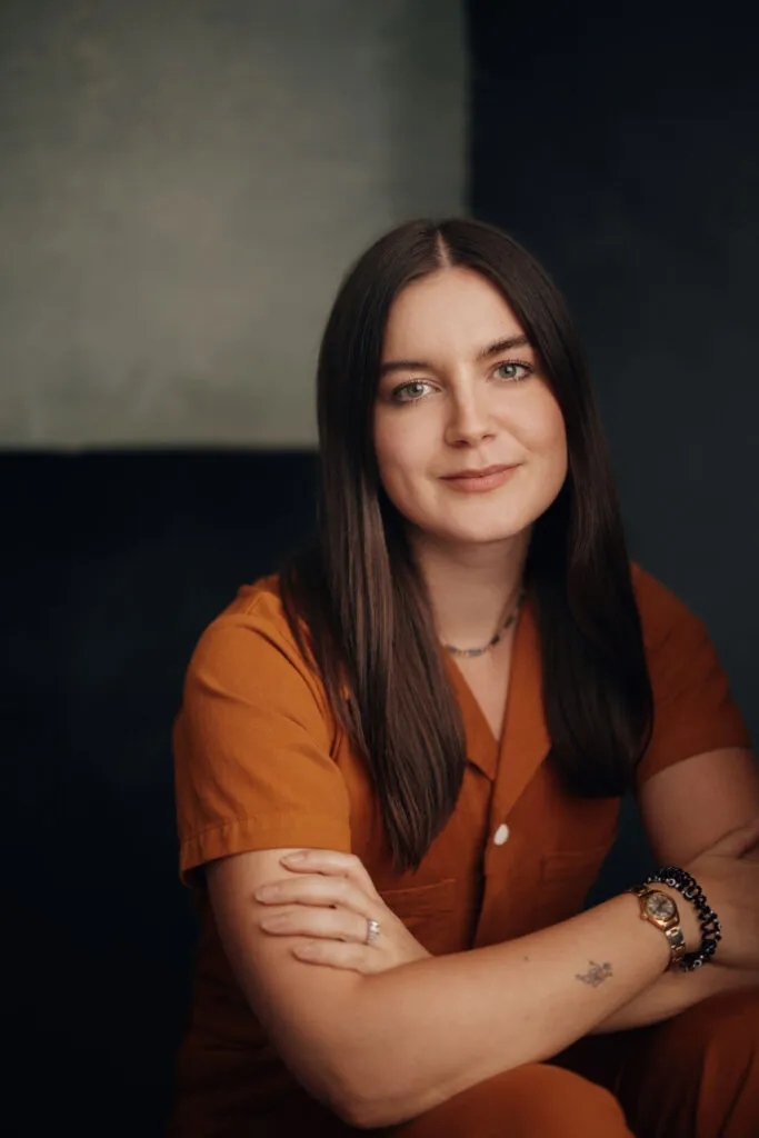 A woman with long brown hair smiles, while sitting forward and leaning her arms on her knees.