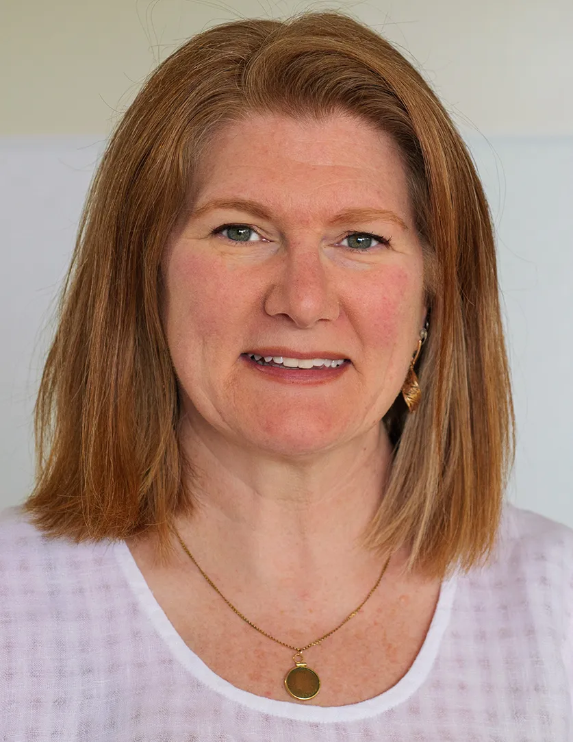 Headshot of a white woman with shoulder-length auburn hair wearing a scoop neck white shirt and a gold necklace
