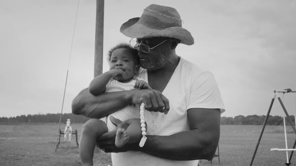 A Black man wearing a straw hat and a white shirt holds a baby girl in his arms.