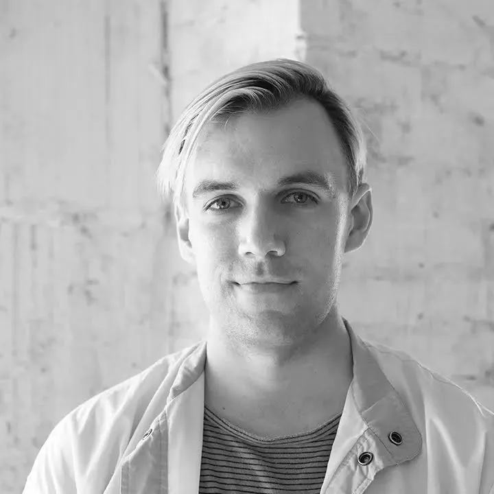 Black and white headshot of a young white man with medium-length dyed blond hair, wearing a striped tee under a jacket.