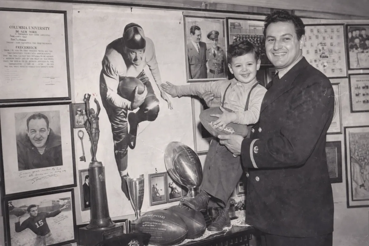 Black and white photo of Sid holding his son Bobby. They are in front of a poster of Bobby as a quarterback.