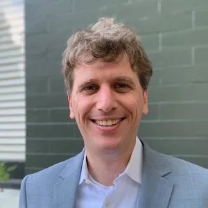 Headshot of a smiling adult man in a white shirt and grey suit in front of a brick wall