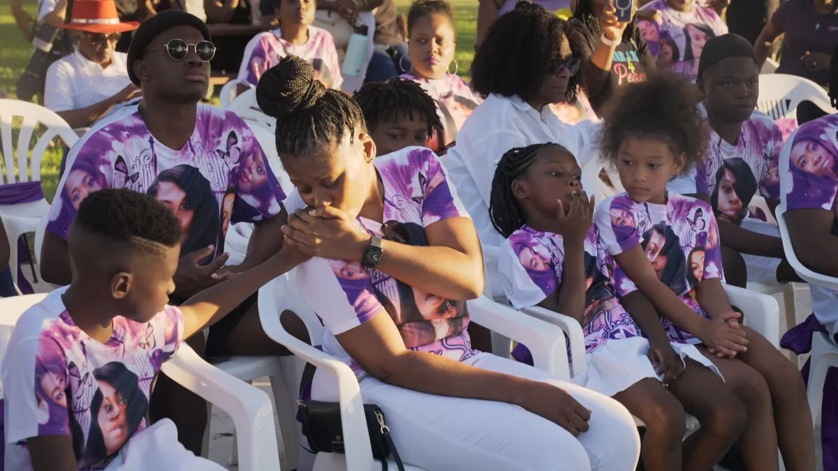 A group of Black boys and girls all wearing a white and purple t-shirt emblazoned with images of a Black woman, sit at an outdoor gathering.