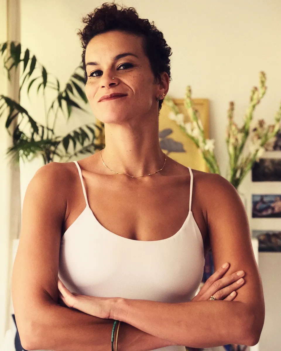 A light-skinned Black woman with short-cropped dark hair, smiles at the camera with her arms crossed while wearing a light tank top