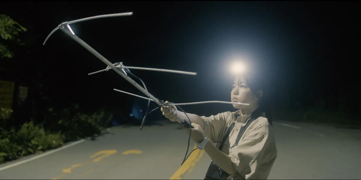 A young Asian woman stands in the middle of the street at night holding an antenna, lit only by the headlamp she sports on her head
