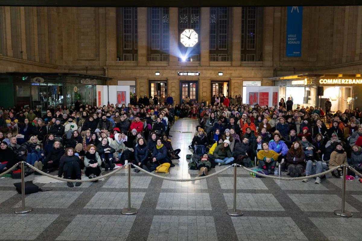 Hundreds of folks sit on the ground at Leipzig's Central Station, at a film screening; a projector can be seen behind them below a clock