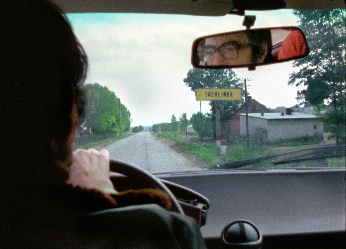View from inside a car, a white man with dark hair and glasses can be seen driving through the rearview mirror as he passes a sign that reads Treblinka