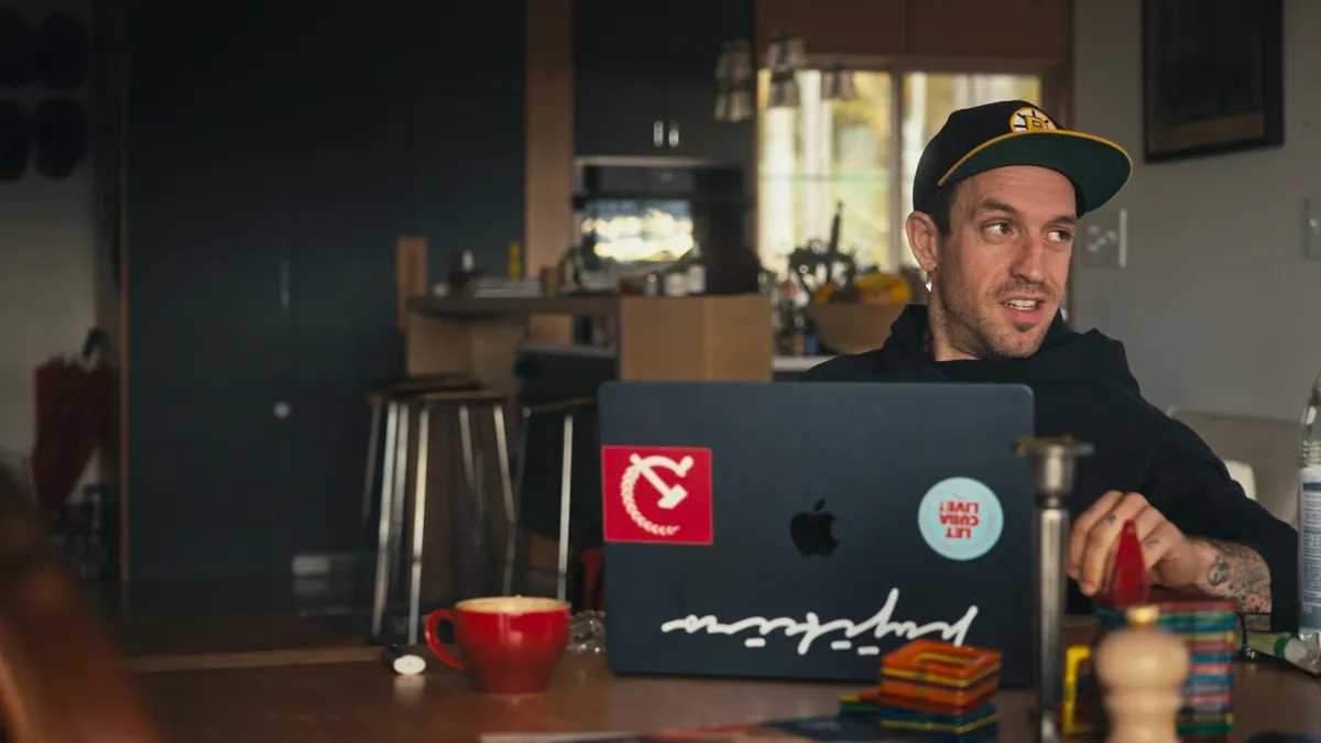 A white man wearing a cap and a hoodie sits at a kitchen table with his laptop, which is adorned by a communism sticker and another that reads "Let Cuba Live"