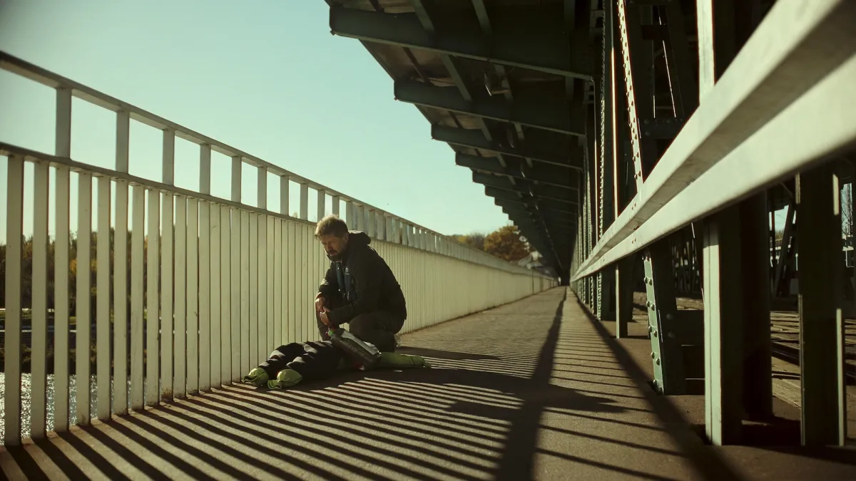 Wide shot of a bearded middle-aged white man with short-cropped hair kneeling over a mannequin of sorts at the railing on a bridge