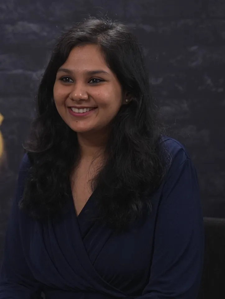 Headshot of a smiling young Brown woman with long dark straight hair, wearing a dark blue shirt in front of a darkened wall
