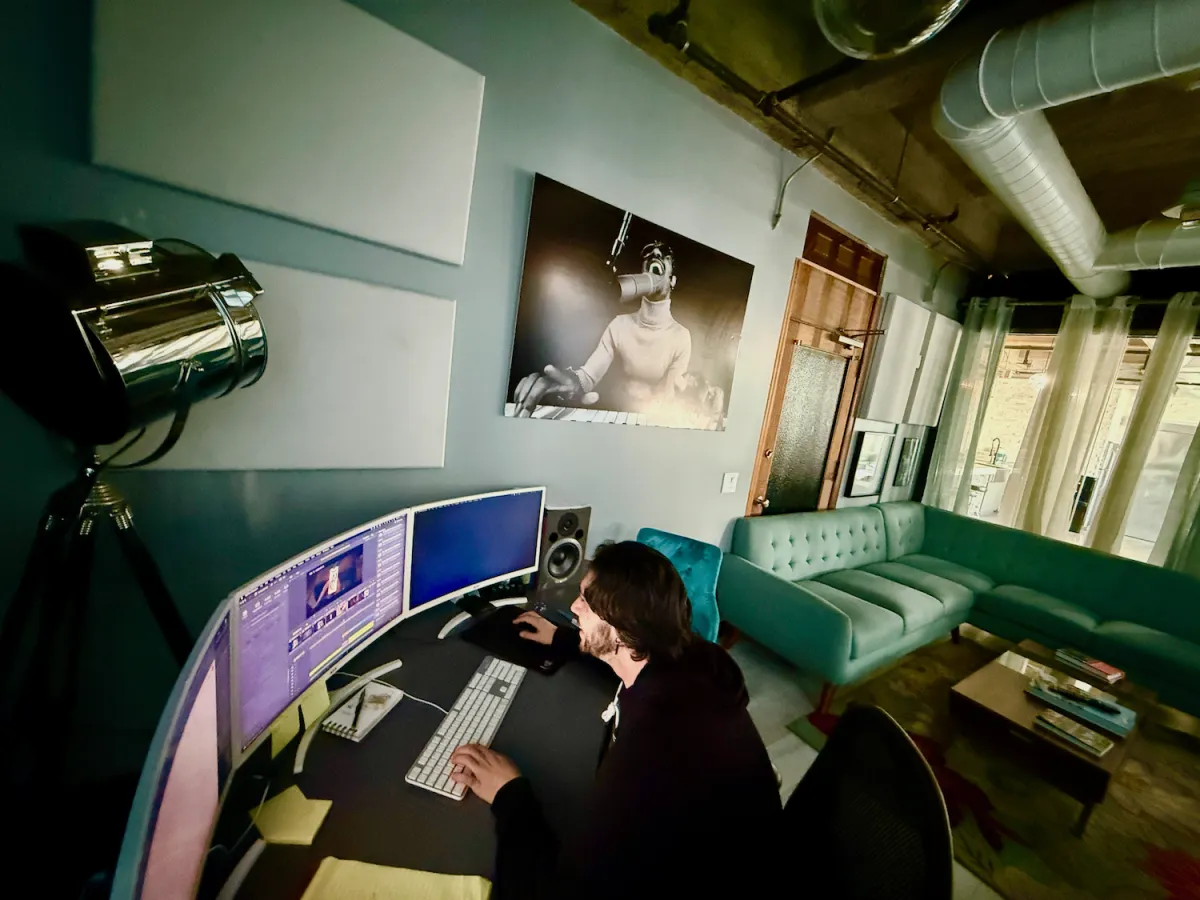 A white man with long black hair in a hoodie sits at an editing bay in a stylish, industrial-looking, emerald-decorated room