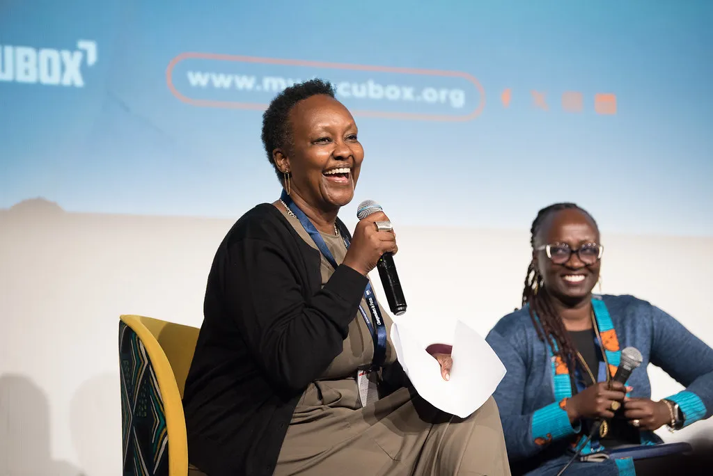 A Black woman with short-cropped hair sits at a stage next to another Black woman; both are holding mics and smiling widely