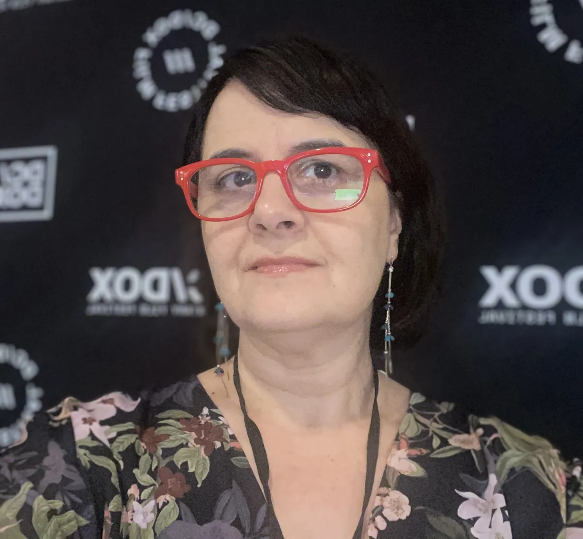 Headshot of a female wearing red-framed glasses and long dangling earrings stands in front of a black step-and-repeat backdrop, dressed in a dark floral top.