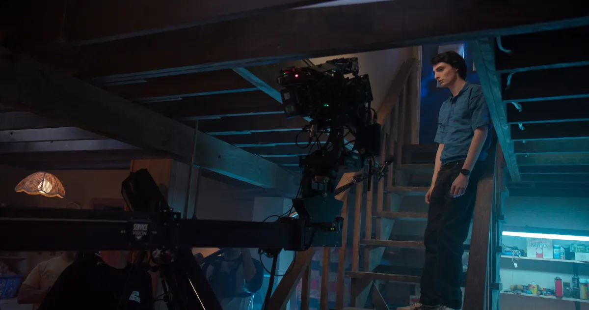 A young white actor wearing a blue button down shirt and pants stands on a basement with a camera rigged in front of him
