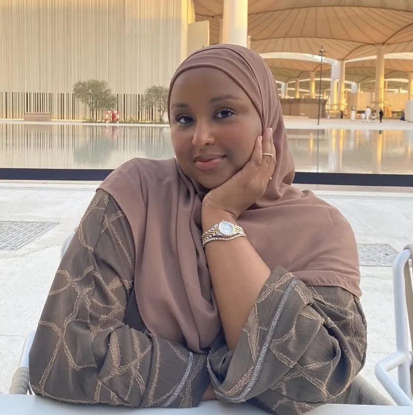 A brown-skinned woman in a dusky pink headcovering sits at an outdoor table.