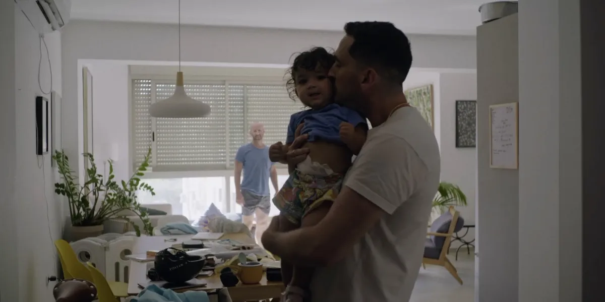 A young brown-skinned man hugs and kisses a young toddler girl amid a slightly messy living room while a balding white man in shirt and shorts looks at them in the background