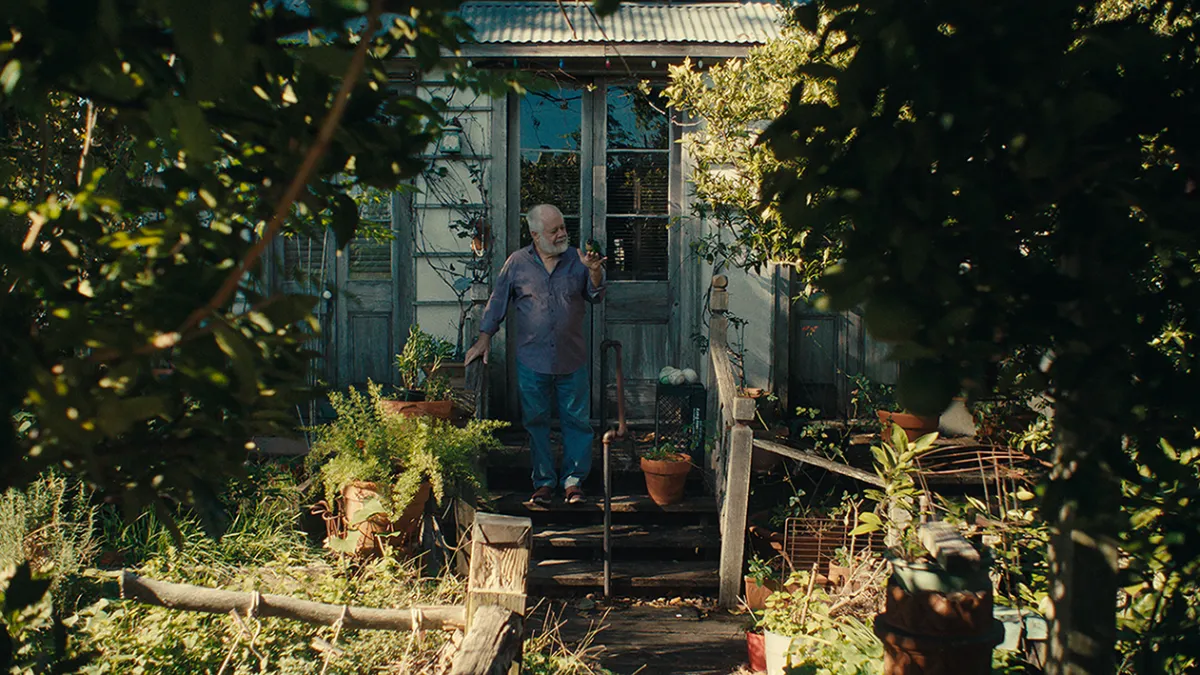 A man in blue jeans and a long sleaved shirt stands in front of the door of his house and his lush, green front yard.